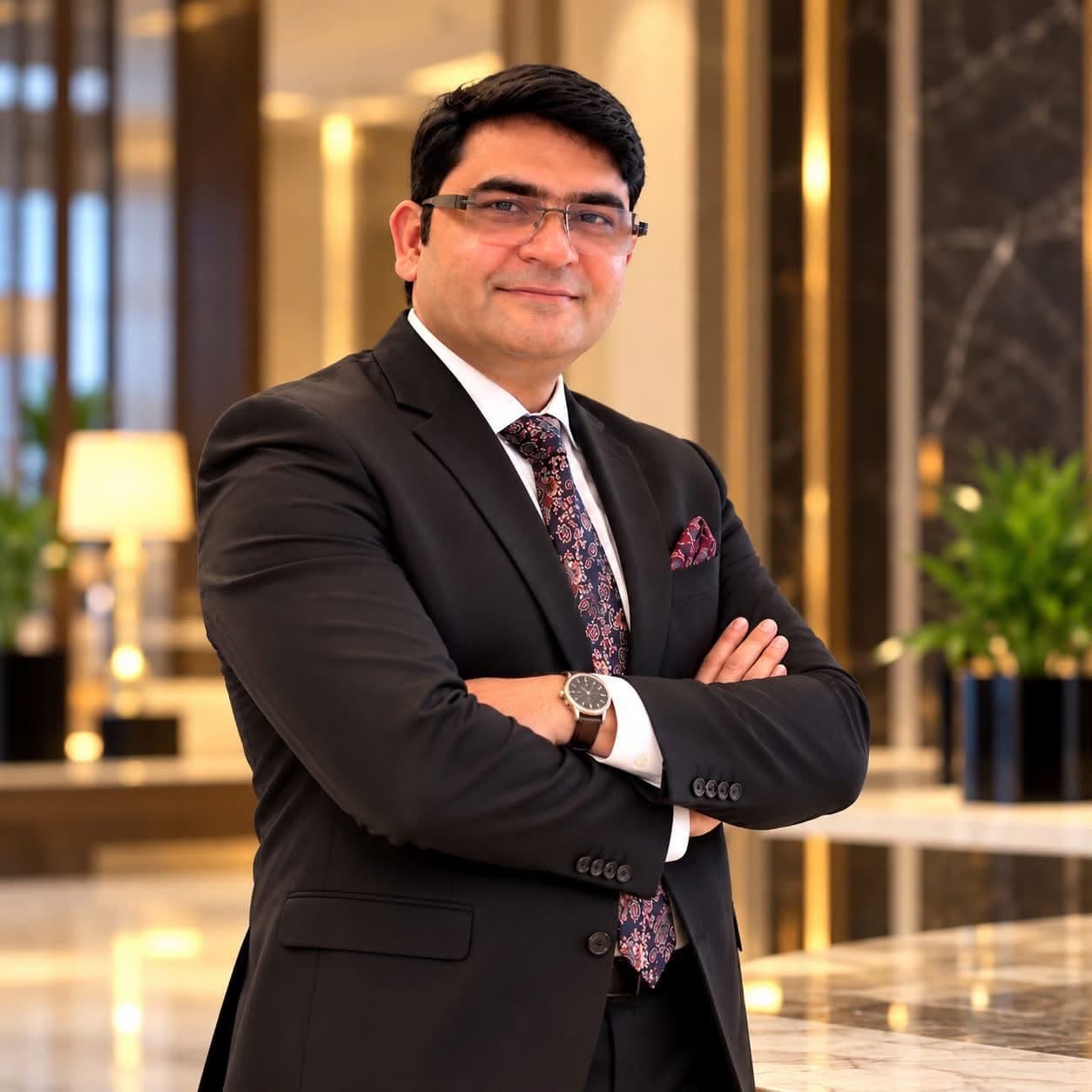 Aadesh Sharma in a dark suit, arms crossed, in a hotel lobby